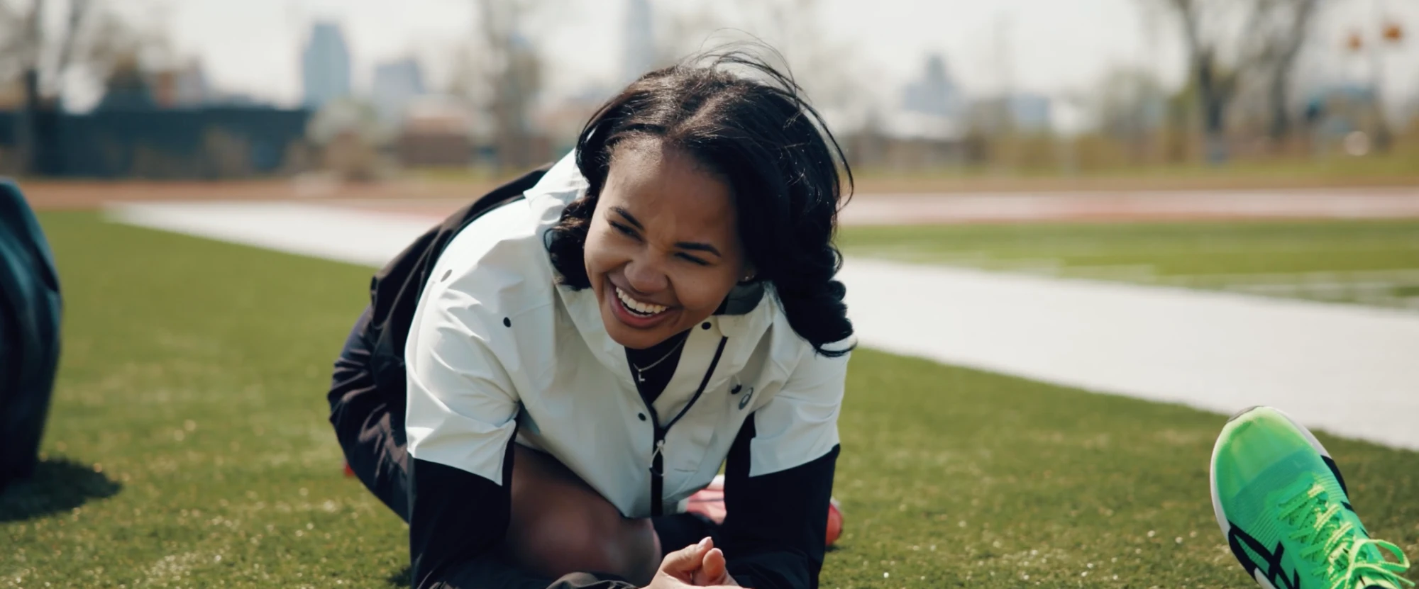 A runner laughs while stretching on the grass beside a track