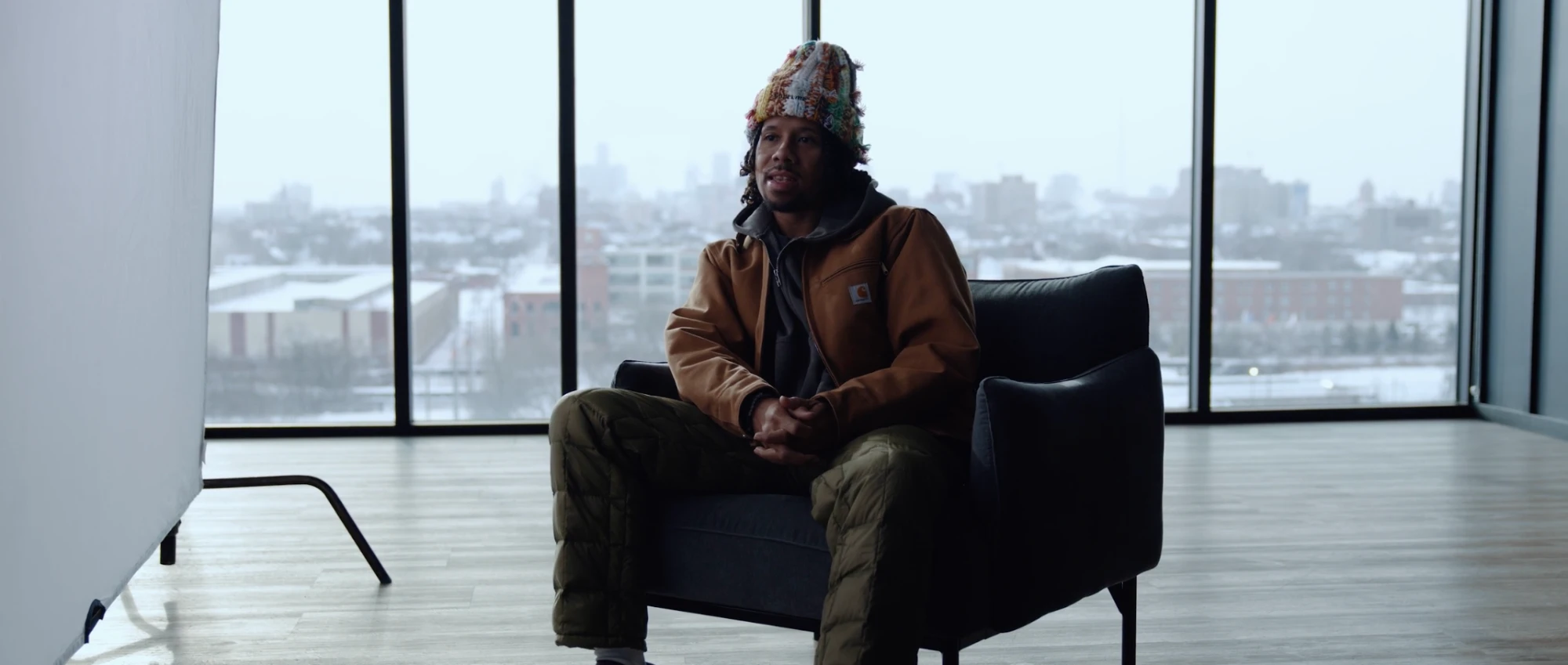 A man in a knit hat and work jacket speaks in front of a wintry city skyline