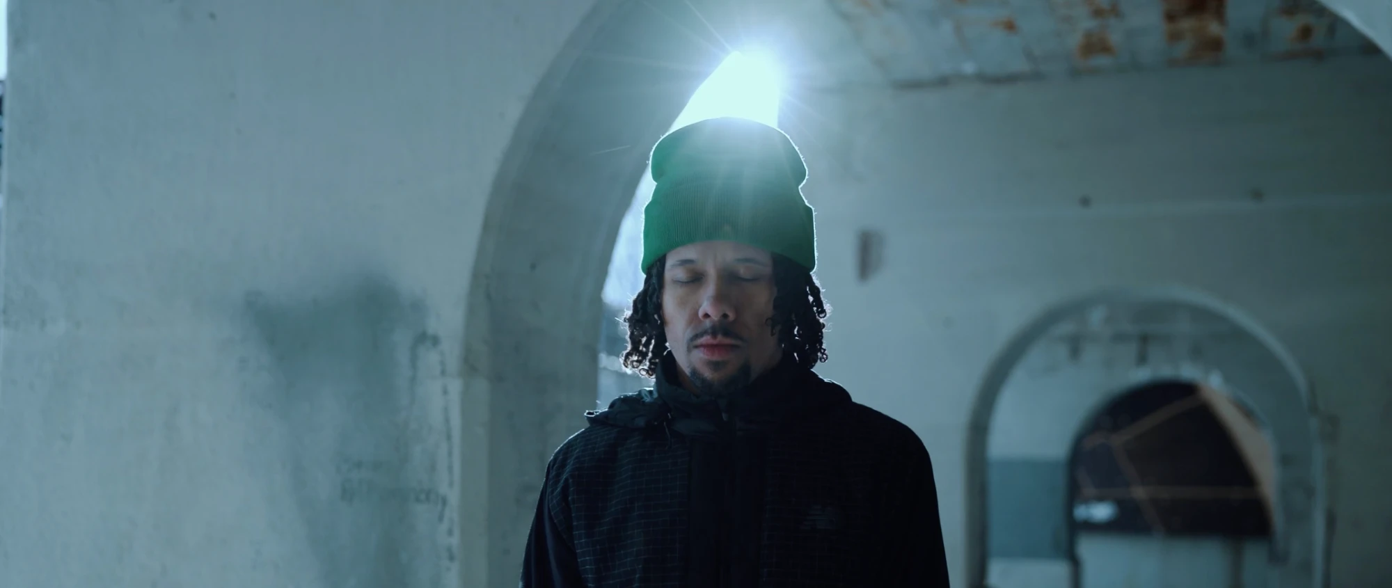 A man in a green beanie stands with eyes closed under a sunlit archway