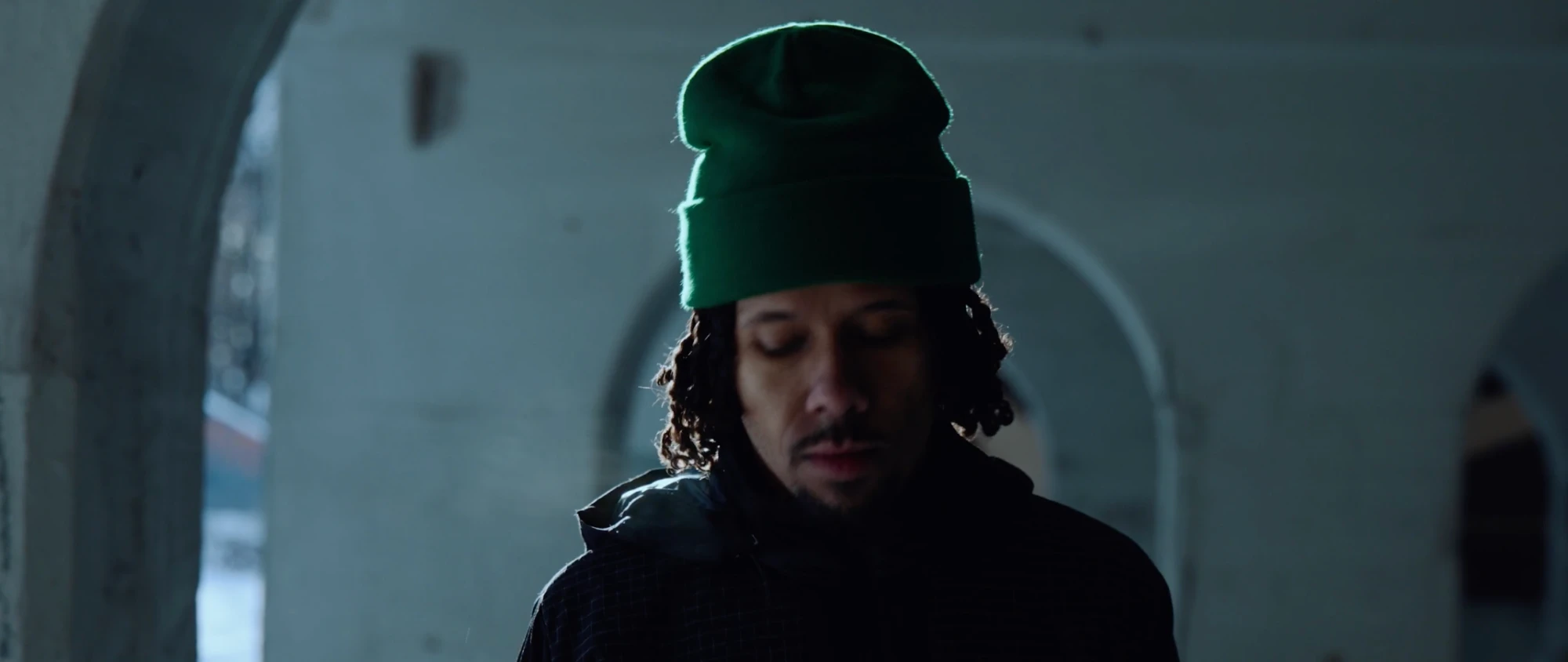 A man in a green beanie looks down, backlit in a concrete arcade