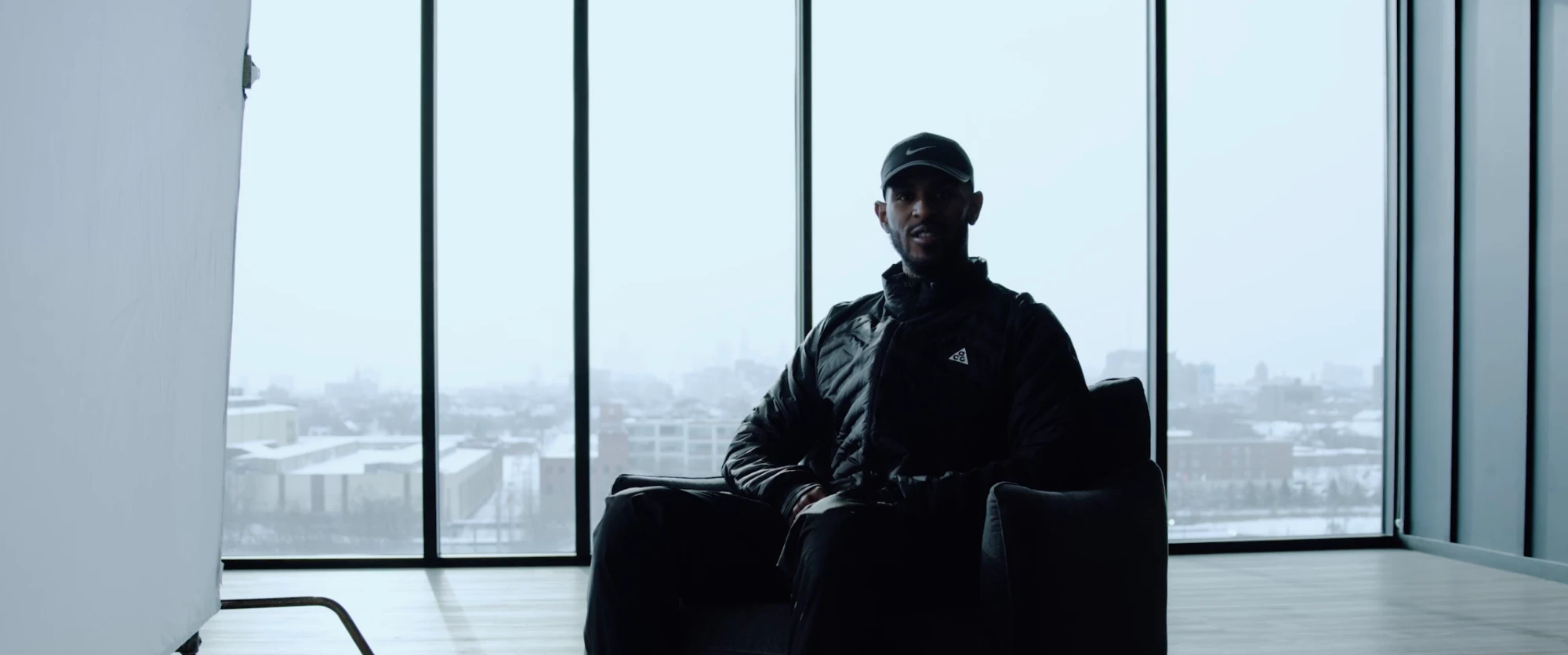 A man in a cap and black jacket sits silhouetted against tall windows